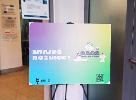 zdjęcie przedstawia sztalugę ze zdjęciem, na którym widnieje napis ZNAJDŹ RÓŻNICĘ oraz grafika przedstawiająca młode osoby z telefonami i laptopami, pośrodku grafiki znajduje się napis GŁOS POKOLENIA 3.0