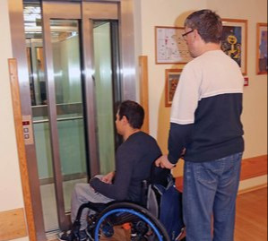 Zdjęcie ilustracyjne do treści artykułu: osoba z niepełnosprawnością poruszająca się na wózku inwalidzkim, prowadzona przez asystenta.