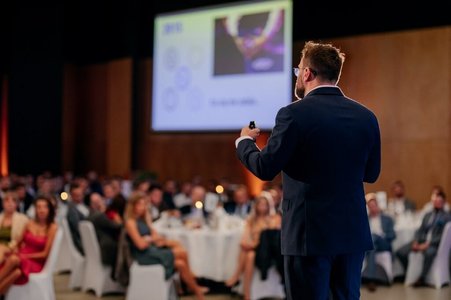 Zdjęcie przedstawia mężczyznę w garniturze trzymającego w lewej ręce mikrofon. W tle widać ludzi ubranych w stroje wieczorowe, siedzących przy stołach. 