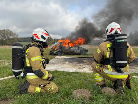 Strażacy w trakcie akcji gaśniczej płonącego pojazdu z wykorzystaniem otrzymanej płachty gaśniczej.