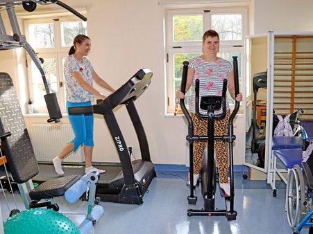 Na zdjęciu dwie kobiety w trakcie rehabilitacji, otoczone różnorodnym sprzętem treningowym.