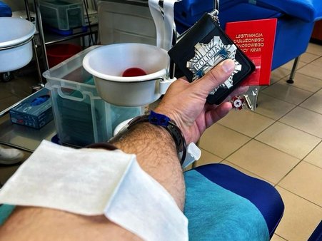 Zdjęcie przedstawiające rękę funkcjonariusza policji, w trakcie honorowego oddawania krwi. W dłoni trzyma odznakę policyjną oraz legitymację dawcy krwi.