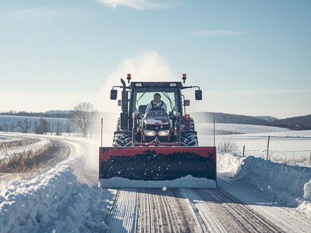 Zdjęcie przedstawia pług odśnieżający lokalną drogę, w tle pola, drzewa i dużo śniegu
