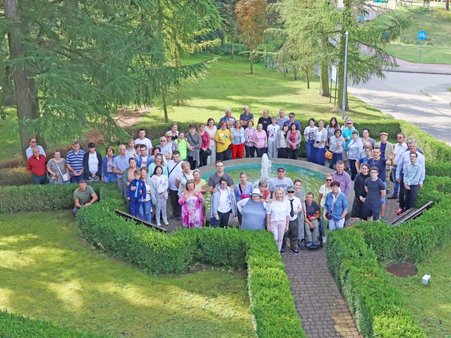 Zdjęcie przedstawia grupę ludzi pozujących do zdjęcia, stojących wokół fontanny, w otoczeniu drzew i żywopłotu