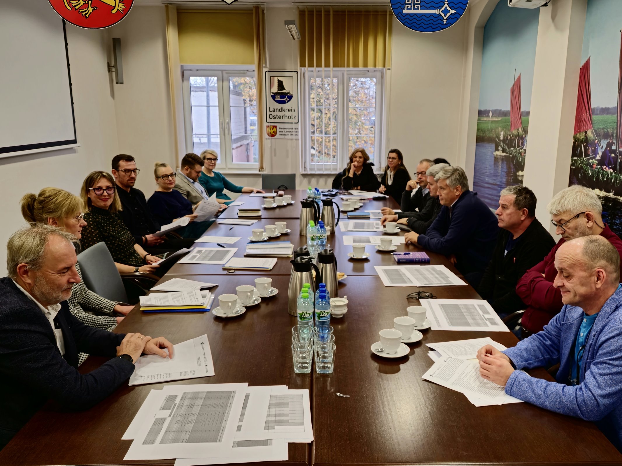 16 członków zespołu siedzi przy stole w sali narad. Zapoznają się z dokumentami projektowymi leżącymi na stole oraz omawiają sprawy istotne dla realizacji programu unijnego