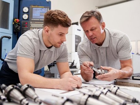 zdjęcie ilustracyjne do artykułu przedstawiające przestrzeń warsztatową na maszynami w tle oraz instruktora demonstrującego stażyście produkt/część metalową 
