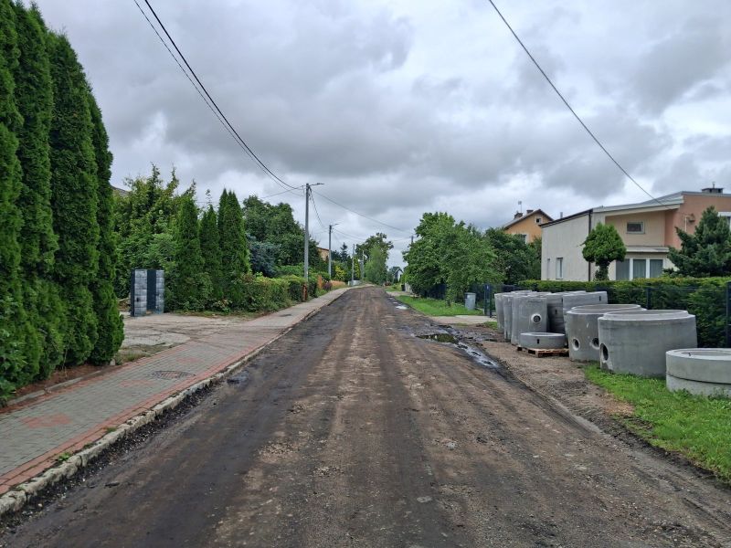 Fragment modernizowanej drogi z zerwaną starą nawierzchnią. Widoczne nierówne podłoże i przygotowania do dalszych prac. W tle drzewa i zabudowania.