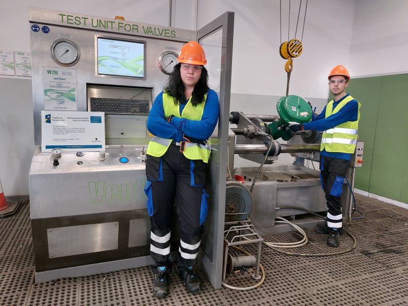 zdjęcie przedstawia stażystów w odzieży roboczej podczas stażu uczniowskiego. Obok nich znajduje się plakat z informacją o dofinansowaniu projektu z Unii Europejskiej